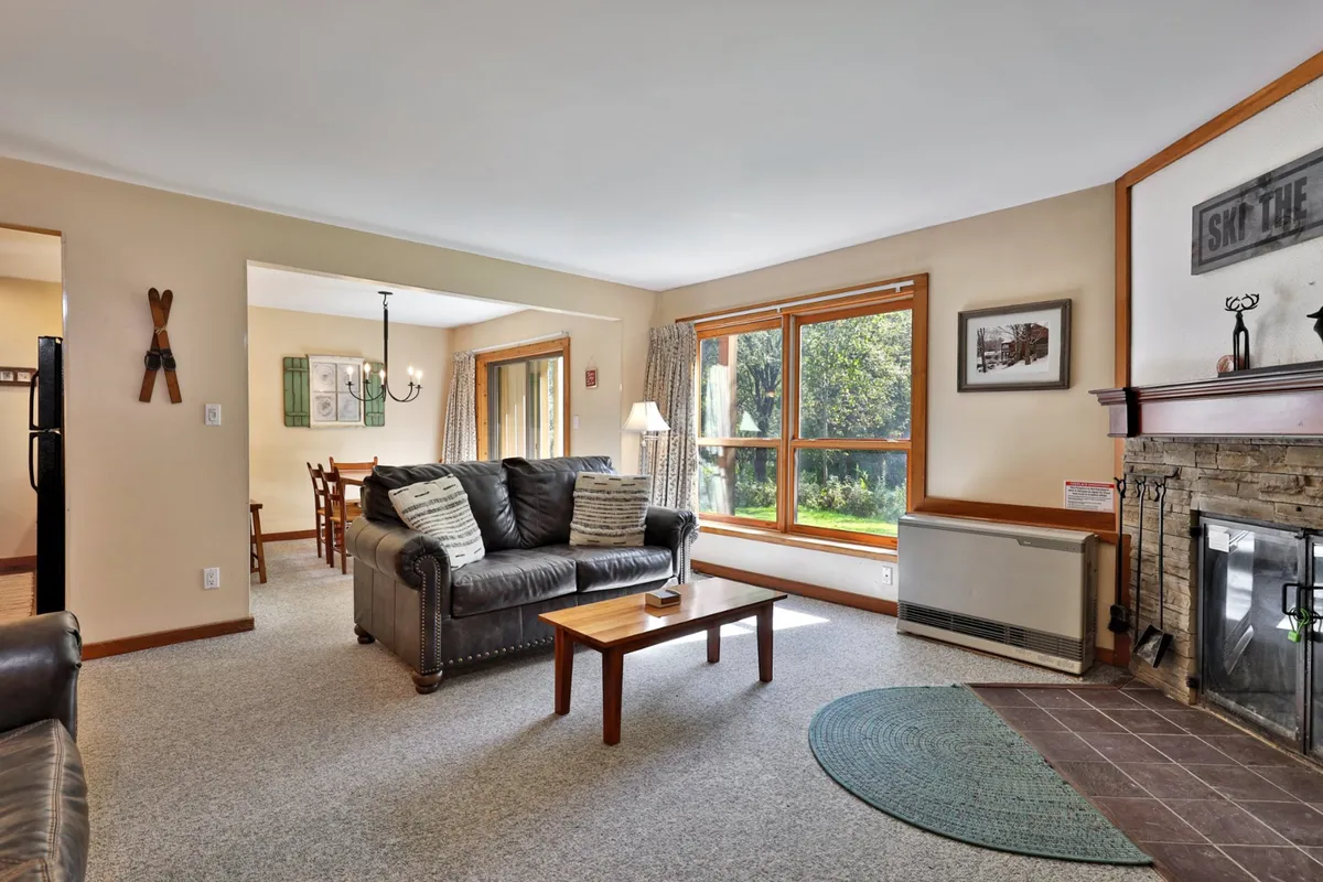 A spacious living room with a large leather sofa, stone fireplace, and generous windows that bathe the space in natural light. In the background, the dining area is visible, hinting at the open-concept layout and flow between communal spaces.