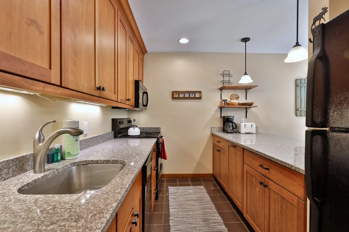 A view of the kitchen showcasing granite countertops, wooden cabinetry, modern appliances, and hanging light fixtures. The coffee station is visible on the counter, and the space radiates a cozy, inviting atmosphere ideal for meal preparation and morning routines.