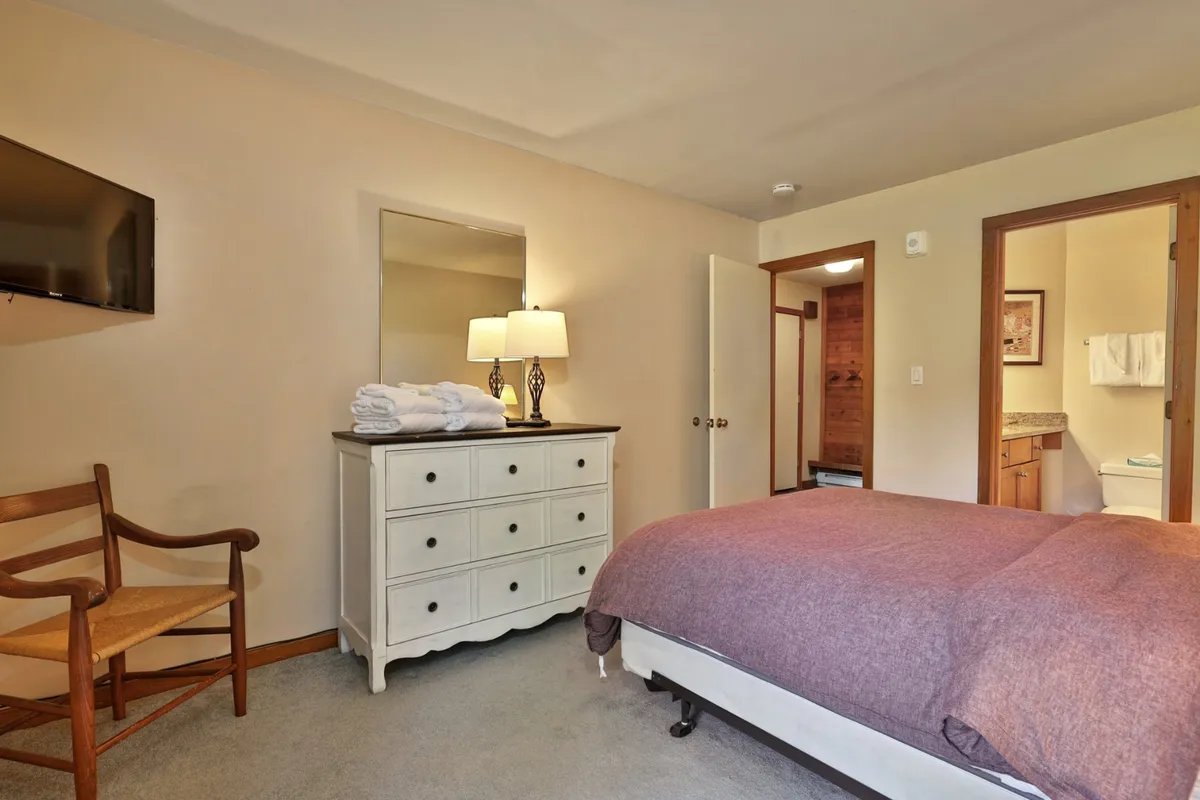 A serene and comfortable bedroom scene featuring a queen bed with a purple blanket, dresser with neatly arranged towels, a mirror, and a lamp, evoking a sense of unwinding and private comfort.