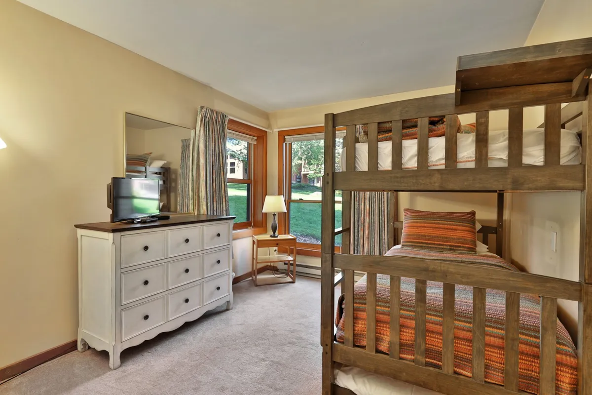 A cozy bedroom featuring a sturdy wooden bunk bed, a dresser, mirror, and a sunlit window. The inviting space is ideal for kids or groups, providing a playful yet restful environment for bedtime stories or games.