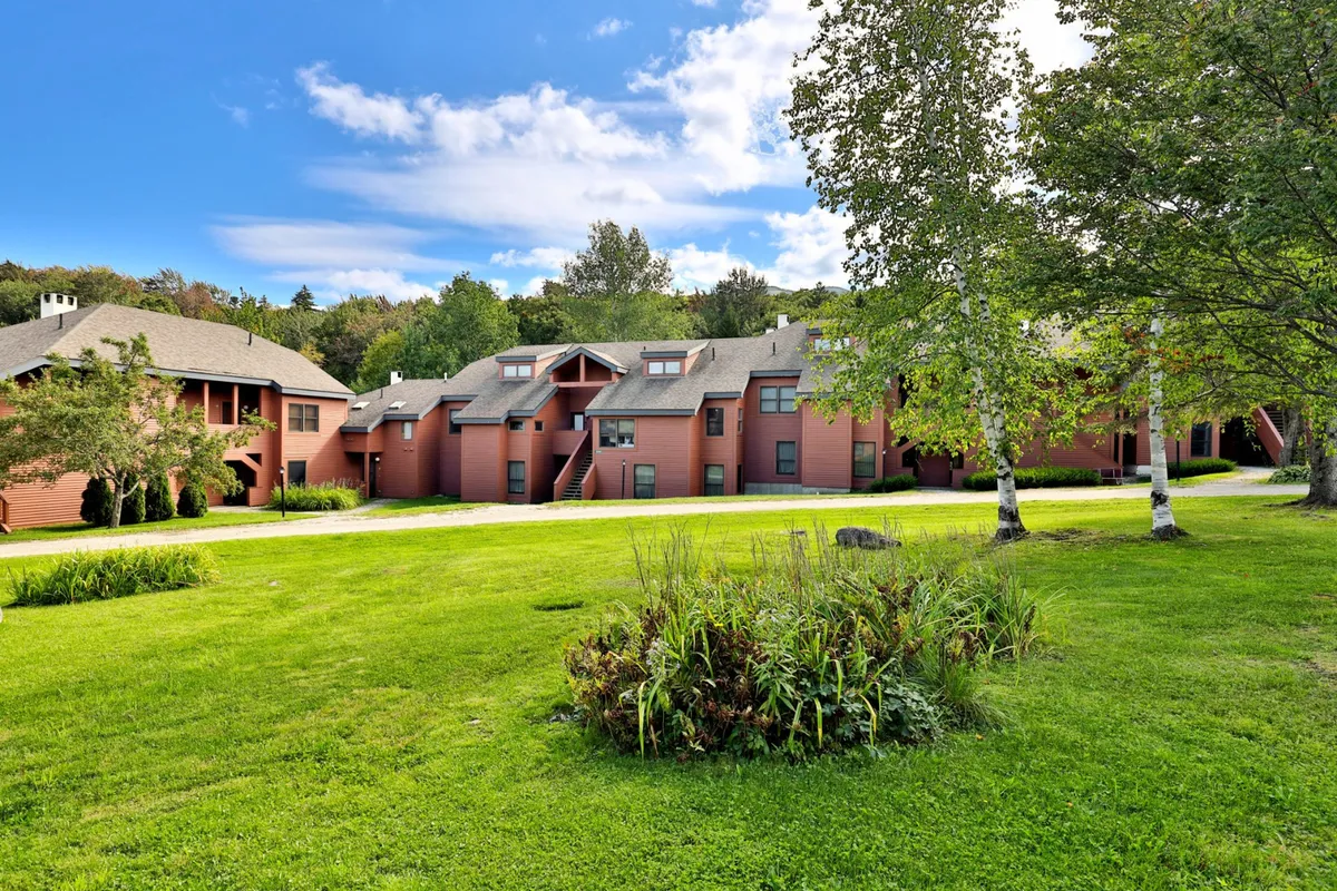 This exterior shot features the red wooden Trail Creek condo buildings set against a lush green lawn and framed by mature trees beneath a clear blue sky. The peaceful scene suggests easy access to outdoors and proximity to mountain activities.