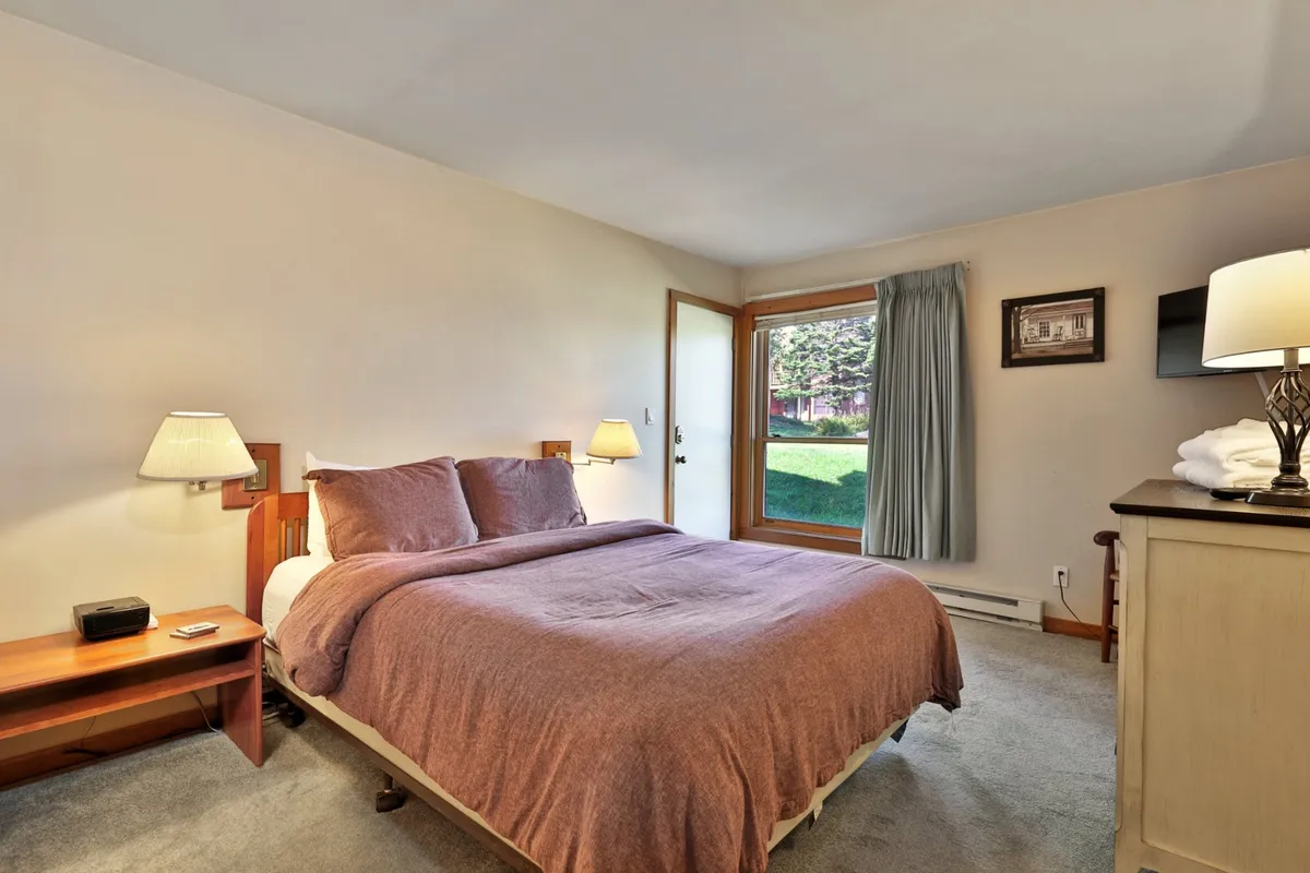 This image displays a serene bedroom featuring a comfortable bed with crisp linens, a window offering nature views, a nightstand, and a soft lamp, creating a peaceful and inviting sleeping environment.