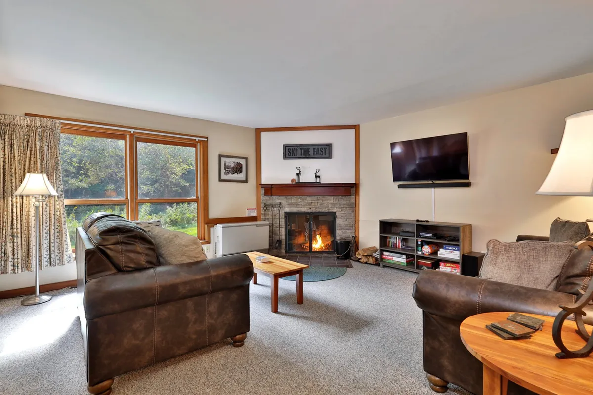 A warm, inviting condo living room with a glowing fireplace, cozy sofa, large windows framing snowy views, and a coffee table perfect for gathering around. The ambiance exudes comfort and relaxation, ideal for unwinding after a day on the slopes.