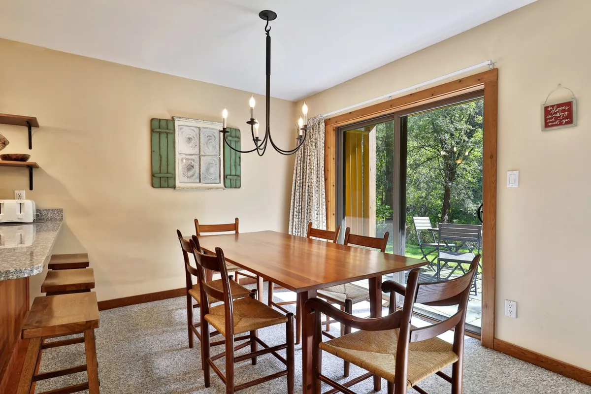 A warm, welcoming dining area featuring a large table under a chandelier, with a glass door providing views of the outdoors. The space suggests shared meals, special occasions, and an easy flow between indoor comfort and outdoor scenery.