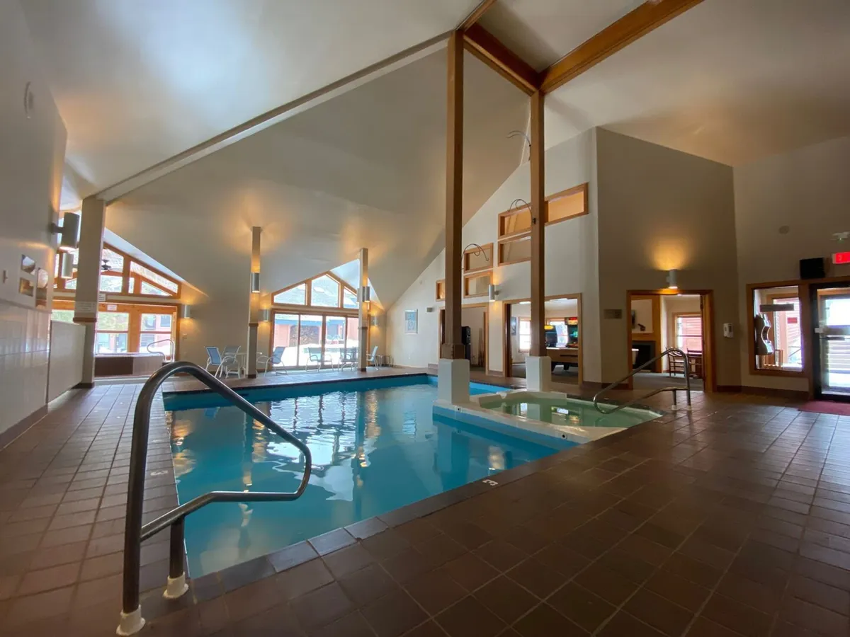 An inviting indoor swimming pool and jacuzzi area, bathed in natural light from large windows and featuring wooden beams. The atmosphere is serene, suggesting a relaxing retreat after a day on the slopes.
