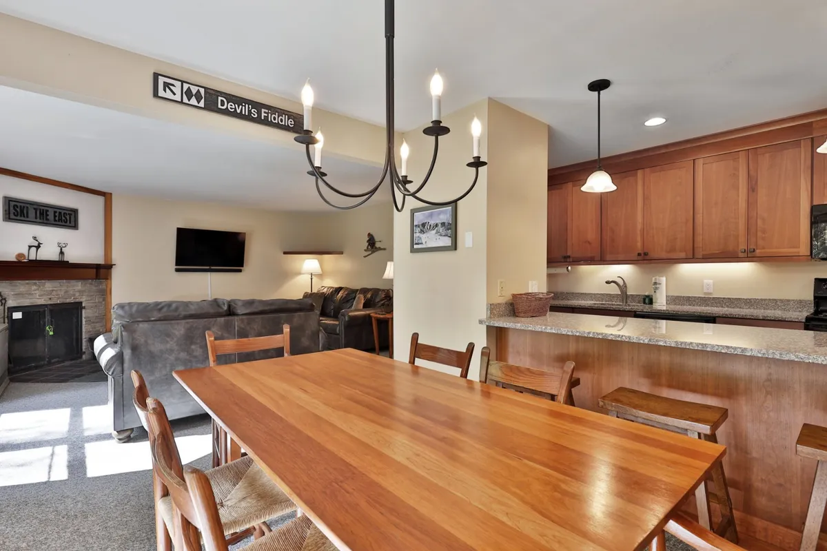 An open-plan dining area with a large dining table under a chandelier, the kitchen counter in the background, and a glimpse of the living area with a sofa and fireplace. The scene suggests gatherings, meals, and a seamless flow between kitchen, dining, and living spaces.