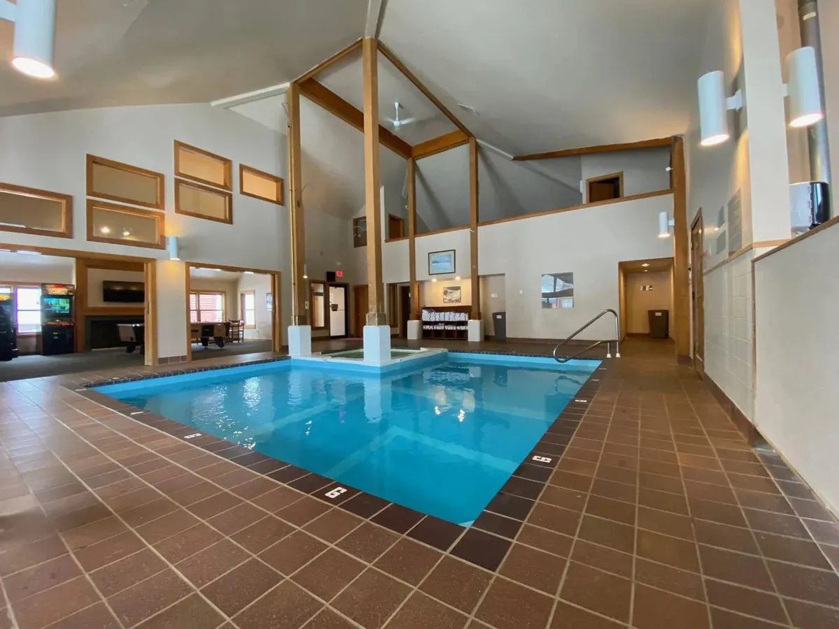 A bright indoor swimming pool area is surrounded by sturdy wooden beams and large windows that let natural light stream in. Comfortable seating along the side invites relaxation and socializing, creating a vibrant and family-friendly atmosphere.
