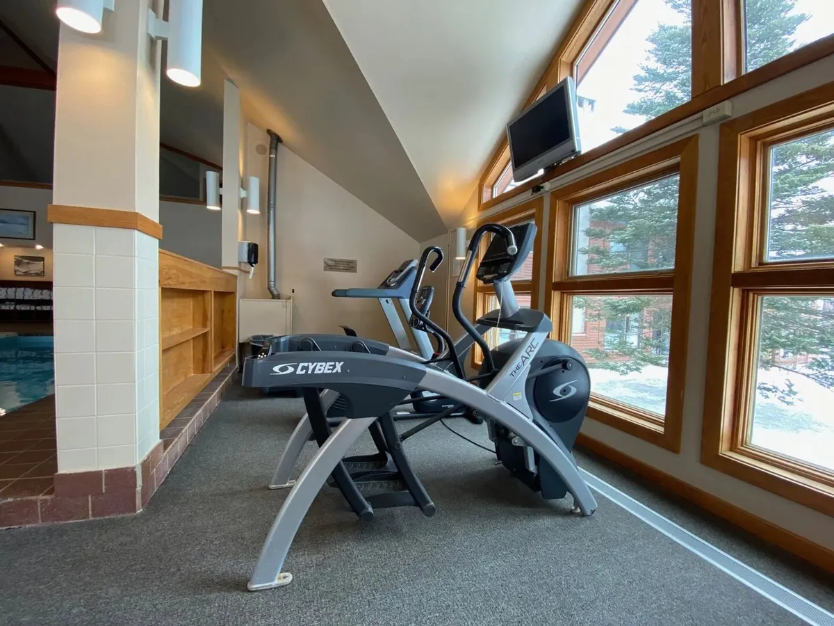 A well-lit fitness room with elliptical machines, large windows offering views, and a TV. The space is designed for active routines and post-exercise relaxation, with plenty of natural light and cues for mountain views beyond.